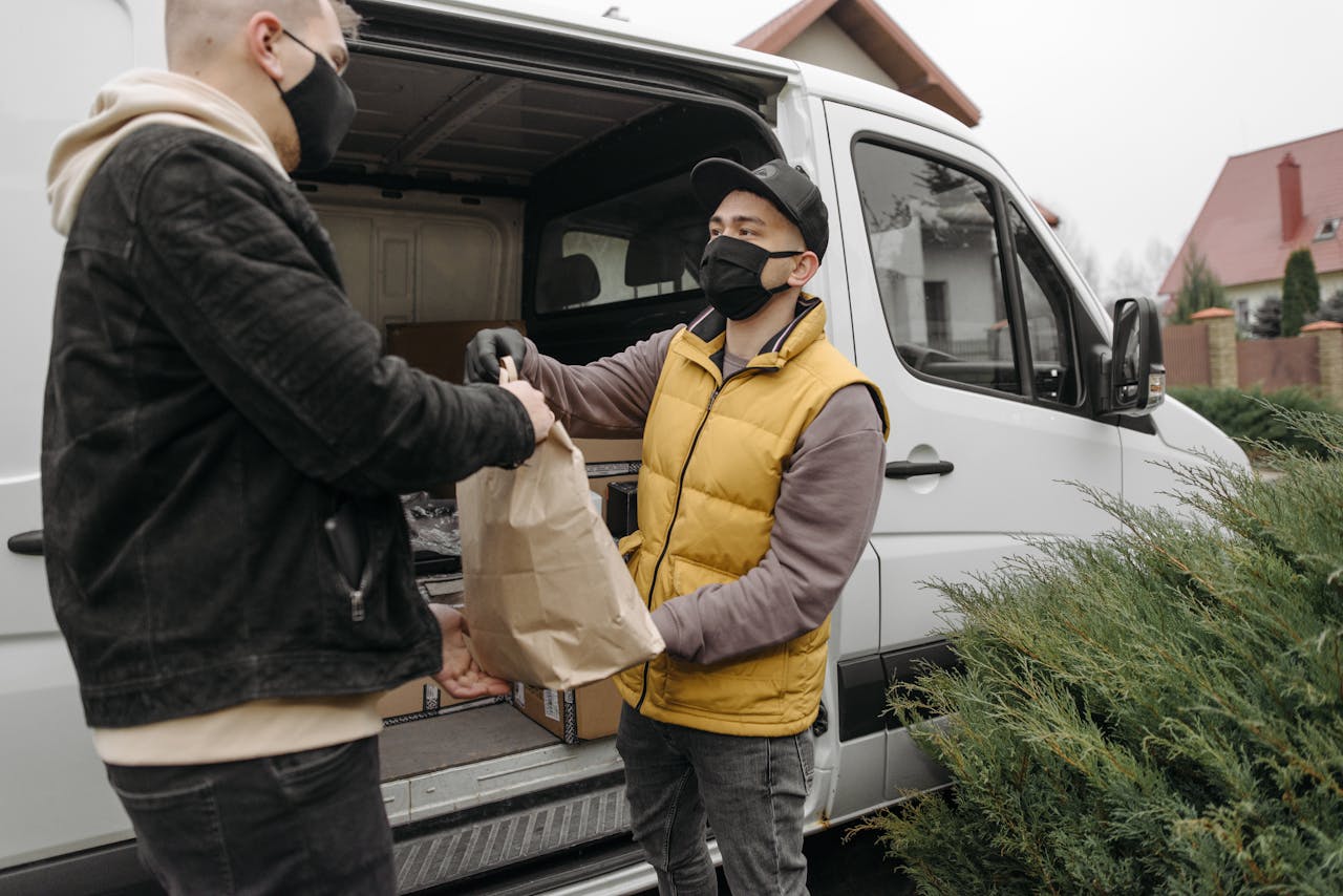 Two men wearing face masks exchange a package by a delivery van outdoors.